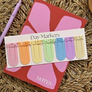 May include: Set of seven colourful plastic bookmark clips with the days of the week spelled out. The clips are arranged in a row on a white background with the words "Day Markers" above them.