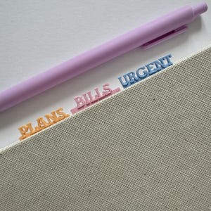 May include: A light purple retractable pen rests above three small, raised plastic tabs attached to a textured beige planner. The tabs are labelled in capital letters: orange for PLANS, pink for BILLS, and blue for URGENT, organised in a row.