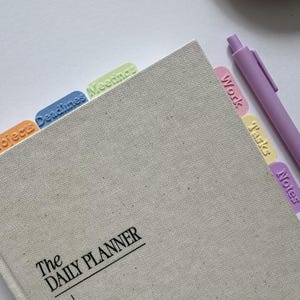 Puede incluir: Una agenda diaria con cubierta de tela beige y el texto The DAILY PLANNER impreso en negro. Incluye pestañas de índice de plástico en colores pastel etiquetadas como Projects, Deadlines, Meetings, Work, Tasks y Notes, junto a un bolígrafo morado claro a juego.