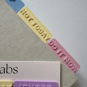 May include: A set of pastel-coloured plastic page tabs featuring embossed text. The tabs include the words HELP in blue, NOT TODAY in yellow, DO IT NOW in pink, and IGNORE in purple. The tabs are arranged on a light-coloured card with the text abs.