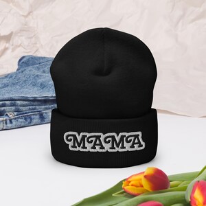May include: Black beanie hat with the word "MAMA" embroidered in white, with a white outline. The hat is made of a soft material and is suitable for cold weather. The hat is a stylish accessory.