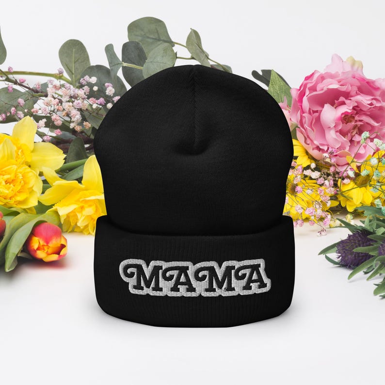 May include: A black beanie hat with the word "MAMA" embroidered in white, surrounded by colorful flowers. The hat is a classic style, suitable for everyday wear.