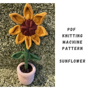 May include: Knitting machine pattern for a sunflower in a pot. The sunflower is yellow with a brown center and green leaves. The pot is light pink.