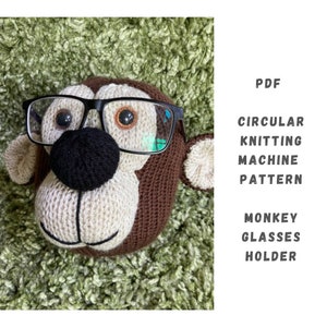 May include: A knitted monkey head with brown fur and a black nose, wearing black framed glasses. The monkey is a glasses holder. The text "PDF CIRCULAR KNITTING MACHINE PATTERN MONKEY GLASSES HOLDER" is in the image.