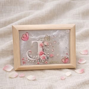 Könnte beinhalten: Ein gerahmtes Kunstwerk mit dem Buchstaben "J", verziert mit rosa Rosen und floralen Akzenten. Das Kunstwerk befindet sich vor einem schimmernden grauen Hintergrund und ist in einem hellen Holzrahmen eingefasst. Rosa Schmetterlings- und Herzdetails ergänzen das Design.