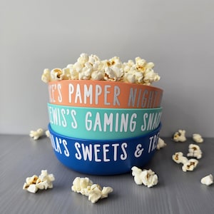 May include: Three brightly coloured bowls stacked on top of each other, filled with popcorn. The bowls are orange, teal, and blue, each with white text. The top bowl says "ME'S PAMPER NIGHT". The middle bowl says "ENIS'S GAMING SNAC".