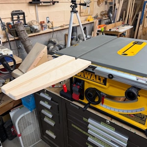 May include: A yellow and black DeWalt table saw featuring a gray metal top and a light-colored wooden extension board attached to the side. The machine includes a black adjustment wheel, a red locking lever, and a printed angle scale on the front.