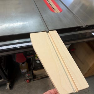 May include: A light-colored wooden table saw push stick with a grooved design. The push stick is held near a table saw with a silver surface and a red and wood blade guard. The saw has a ruler along the side.