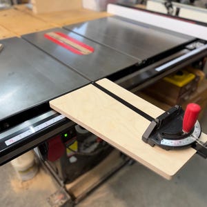 May include: A table saw with a black and silver surface, a red blade guard, and a wooden guide. The saw has a red handle and a measuring scale. A light-colored wooden board is attached to the saw.