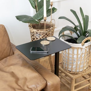 Könnte beinhalten: Ein schwarzer Beistelltisch mit einer rechteckigen Tischplatte und einem einzelnen schwarzen Metallbein. Ein Smartphone liegt auf der Tischplatte. Der Tisch steht neben einem braunen Ledersofa. Im Hintergrund befinden sich zwei Topfpflanzen und ein Kerzenhalter mit zwei weißen Kerzen.