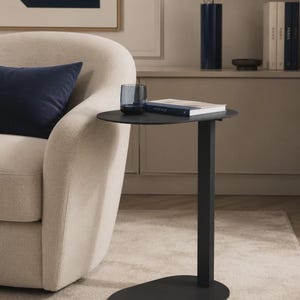 Può includere: Un tavolino a forma di C nero opaco con piano e base ovali, dotato di un'unica colonna di supporto verticale. La superficie del tavolo ospita un bicchiere in vetro blu scuro e un libro con copertina blu scuro, posizionato accanto a una poltrona color crema.