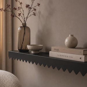 Peut inclure: Une étagère murale noire à bord dentelé supporte un vase en verre brun cannelé avec des branches, un petit bol beige, un vase beige rond et deux livres intitulés Aesop et The Kinfolk Home empilés horizontalement sur la surface plane.