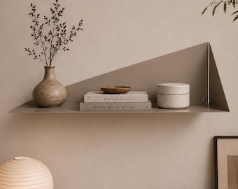 May include: A taupe metal wall shelf with a triangular side panel holding a textured vase with branches, a small wooden bowl, two stacked books titled Kinfolk and Remote Places to Stay, and a cylindrical lidded container in a speckled finish.