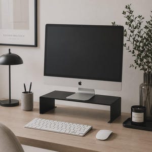 Puede incluir: Un soporte para monitor de metal negro mate con una superficie rectangular y dos soportes laterales, colocado sobre un escritorio de madera clara. El soporte sostiene un monitor plateado, mientras que un teclado y un ratón inalámbricos blancos descansan.