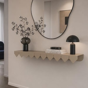 Peut inclure: Une étagère murale flottante beige avec un bord festonné décoratif, comprenant un vase en verre gris foncé avec des branches, deux livres empilés intitulés Kinfolk et Live Beautiful, et une petite lampe noire en forme de champignon sous un miroir.