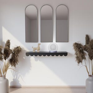 May include: Three arched mirrors above a black scalloped shelf with a wooden dog and a glass orb lamp. The white wall is complemented by tall dried plants in vases, creating a modern, minimalist aesthetic. The shelf is a stylish home decor item.