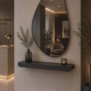 Peut inclure: Un miroir mural asymétrique à cadre noir est suspendu au-dessus d'une étagère flottante noire mate. Sur l'étagère se trouvent un vase en verre foncé contenant des branches feuillues vertes et un petit bougeoir en verre foncé avec une bougie allumée.