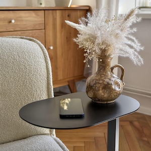 Peut inclure: Une table d'appoint noire de forme ovale avec un smartphone posé dessus. Un vase décoratif avec des fleurs séchées est posé sur la table. Le vase est marron avec un motif unique. Une chaise crème est au premier plan.