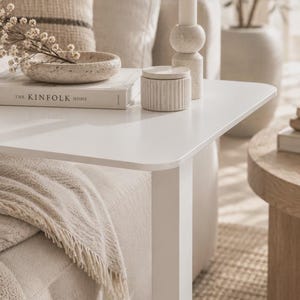 Può includere: Un tavolino bianco a forma di C con un piano rettangolare piatto. Sulla superficie si trovano un libro intitolato The Kinfolk Home, una ciotola in pietra beige maculata con fiori secchi, un piccolo contenitore cilindrico a coste.