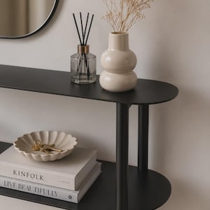 Könnte beinhalten: Ein schwarzer Konsolentisch mit zwei Ebenen und abgerundeten Kanten. Auf der oberen Ablage stehen ein durchsichtiger Glas-Diffusor mit schwarzen Stäbchen und eine cremefarbene Keramikvase mit Trockenblumen. Unten eine cremefarbene Schale mit Schlüsseln.