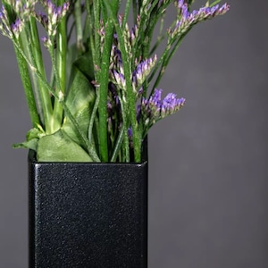 May include: A black square vase with a textured surface holds a bouquet of purple and green flowers.