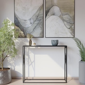 Puede incluir: Una consola de metal negro con una parte superior rectangular y patas geométricas. Dos pinturas abstractas cuelgan sobre la mesa, con colores neutros y líneas curvas. Se exhiben objetos decorativos, incluyendo un jarrón y un cuenco, sobre la mesa.