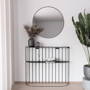 Puede incluir: Una consola moderna de metal negro con un diseño de barras verticales, coronada por un jarrón gris, un pequeño portavelas de cristal y un libro. Un espejo redondo negro cuelga encima. Una planta en maceta está a la derecha.