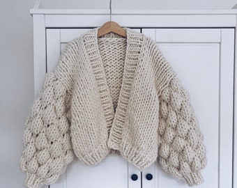 May include: A cream-colored knitted cardigan with a V-neck and large, puffy sleeves. The cardigan is hanging on a wooden hanger against a white background.