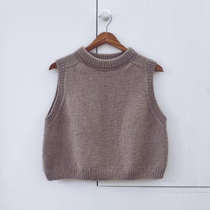 May include: A sleeveless, taupe-colored sweater vest with a ribbed hem and collar, hanging on a wooden hanger. The vest has a high neckline and a simple, classic design, perfect for layering.