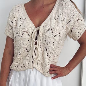 May include: Cream-colored, short-sleeved knit top with a V-neck and tie closure. The top features an openwork pattern and is paired with a white skirt. The knitwear has a delicate, textured appearance, suitable for warm weather.