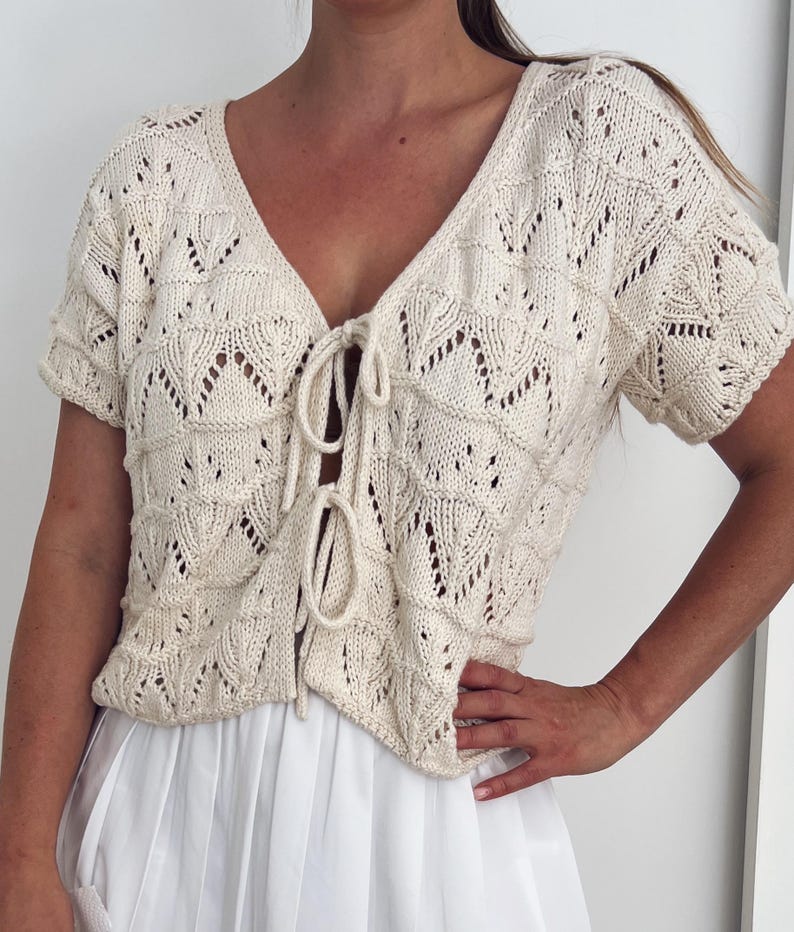 May include: Cream-colored, short-sleeved knit top with a V-neck and tie closure. The top features an openwork pattern and is paired with a white skirt. The knitwear has a delicate, textured appearance, suitable for warm weather.