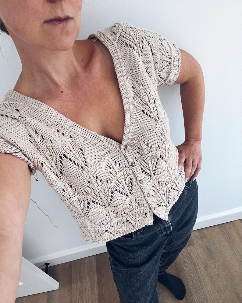 May include: A beige, short-sleeved cardigan with a V-neck and button closure. The knitwear features an openwork leaf pattern. The cardigan is paired with dark wash jeans.