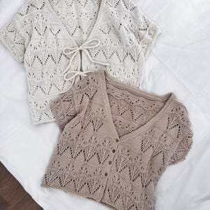 May include: Two short-sleeved, open-front knit cardigans. One is cream-colored with a tie closure, and the other is tan with button closures. Both feature a delicate, openwork leaf pattern, and are displayed on a white surface.
