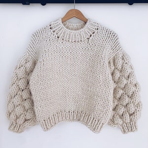 Könnte beinhalten: Ein cremefarbener Strickpullover mit Rundhalsausschnitt und bauschigen Ärmeln.
