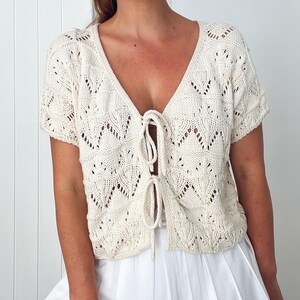 May include: Cream-colored, short-sleeved knit top with a deep V-neck and tie closure. The top features a delicate, openwork leaf pattern throughout. The garment is a light, airy style, suitable for layering or warmer weather.