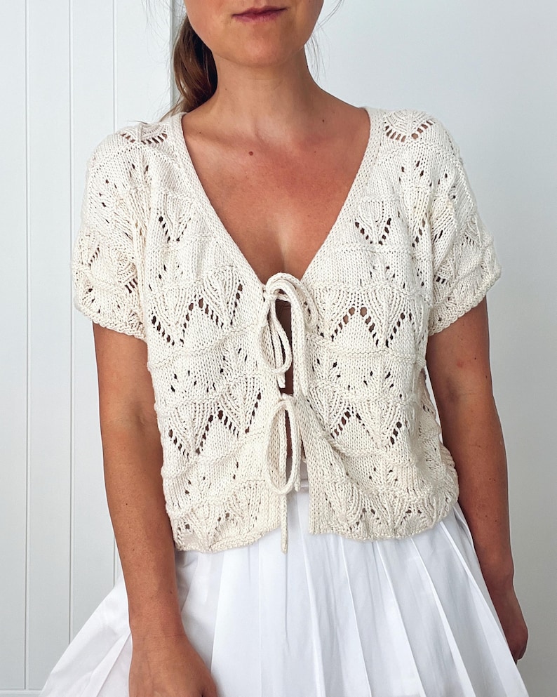 May include: Cream-colored, short-sleeved knit top with a deep V-neck and tie closure. The top features a delicate, openwork leaf pattern throughout. The garment is a light, airy style, suitable for layering or warmer weather.