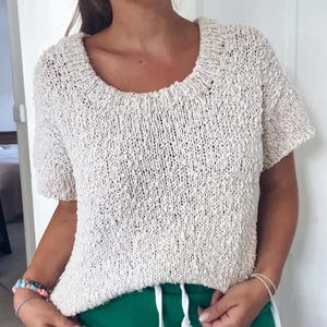 Pode incluir: Um top de malha creme de manga curta com decote redondo largo. O top tem uma aparência texturizada e é combinado com calções verdes. Os calções têm um cordão branco e riscas brancas na lateral.