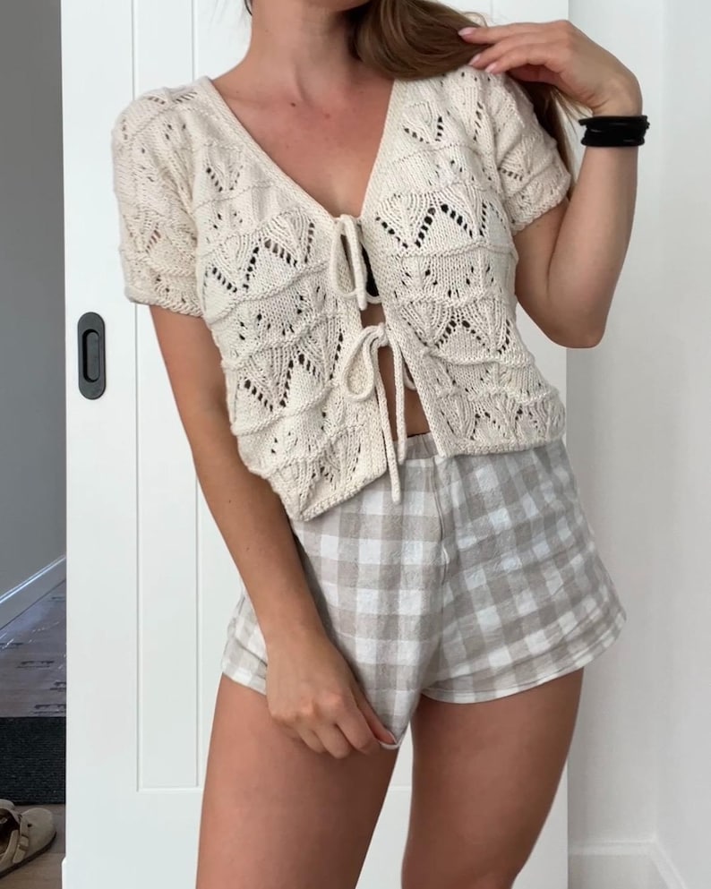May include: Cream-colored, short-sleeved knit cardigan with a V-neck and tie closure. Paired with beige and white checkered shorts. The cardigan features a detailed openwork design. Casual summer outfit.