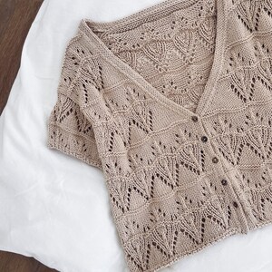 May include: A beige, short-sleeved cardigan with a V-neck and button closure. The knitwear features a delicate openwork pattern with leaf-like designs. The garment is made of a soft, lightweight material, perfect for layering.