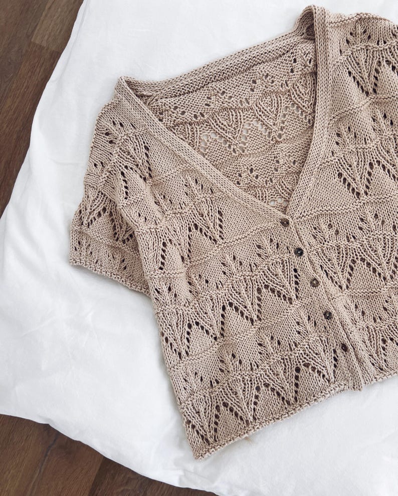 May include: A beige, short-sleeved cardigan with a V-neck and button closure. The knitwear features a delicate openwork pattern with leaf-like designs. The garment is made of a soft, lightweight material, perfect for layering.