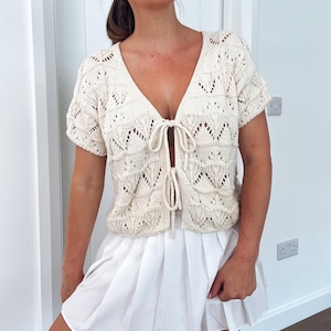May include: Cream-colored, short-sleeved knit top with a deep V-neck and tie closure. The top features a textured, openwork design. Paired with a white pleated skirt. The outfit is suitable for warm weather.