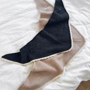 May include: Two triangular knitted scarves, one in dark charcoal grey and one in a light beige tone. Each scarf features a delicate, off-white crocheted lace trim along the edges and a small circular opening near the top point of the fabric.