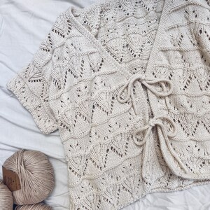 May include: Cream-colored, short-sleeved, open-front cardigan with a textured, open-knit design. The cardigan features a tie closure at the waist and is displayed with several balls of yarn, suggesting a handmade or craft item.