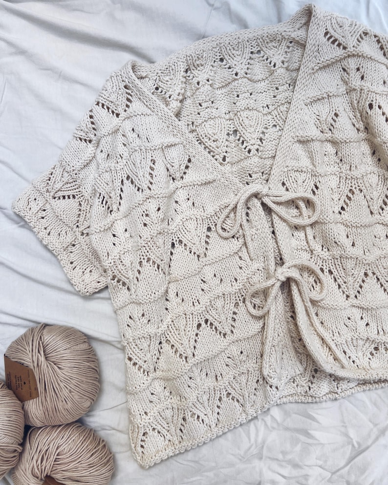 May include: Cream-colored, short-sleeved, open-front cardigan with a textured, open-knit design. The cardigan features a tie closure at the waist and is displayed with several balls of yarn, suggesting a handmade or craft item.