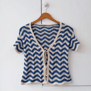 May include: A short-sleeved knit cardigan featuring a blue and cream chevron pattern. The garment has a deep V-neckline, a front opening secured with two thin matching ties, and a solid cream-colored trim along the edges and sleeve cuffs.
