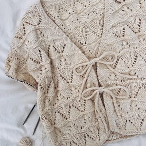 May include: Cream-colored, open-front knitted cardigan with a delicate leaf pattern. The cardigan features short sleeves and a tie closure at the waist. Knitting needles and a ball of yarn are visible, suggesting a handmade item.