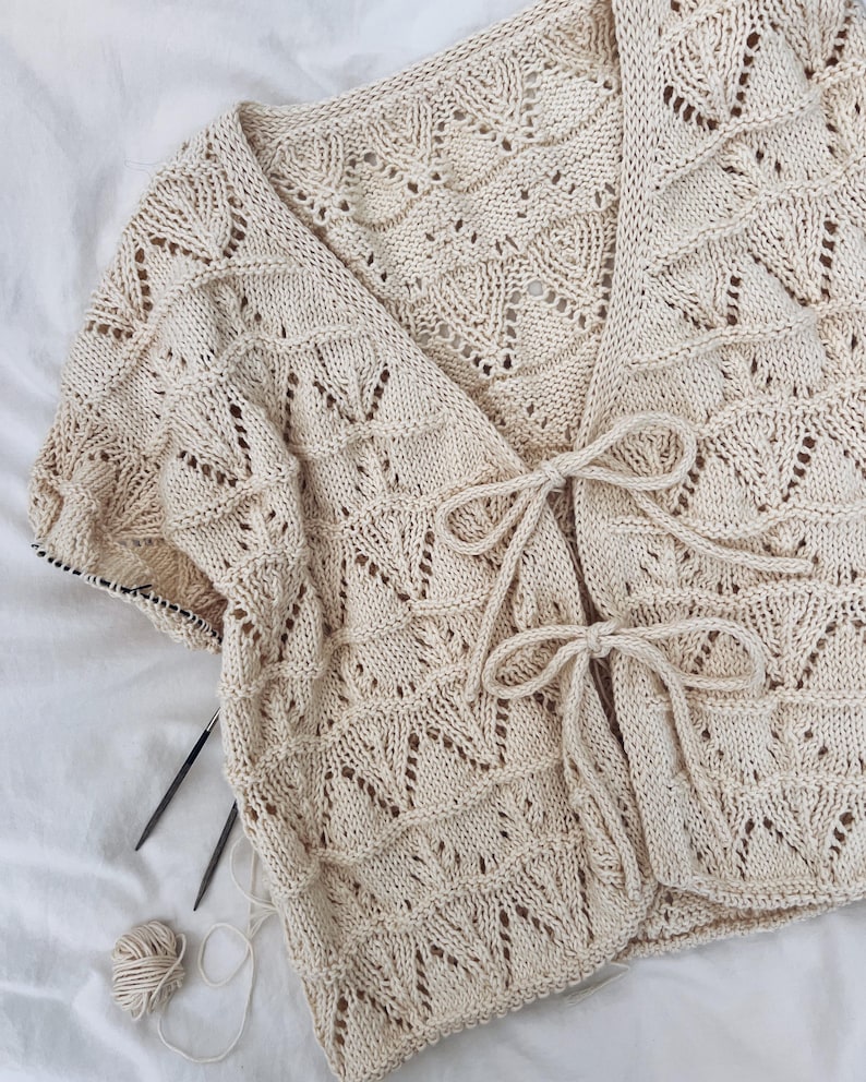 May include: Cream-colored, open-front knitted cardigan with a delicate leaf pattern. The cardigan features short sleeves and a tie closure at the waist. Knitting needles and a ball of yarn are visible, suggesting a handmade item.