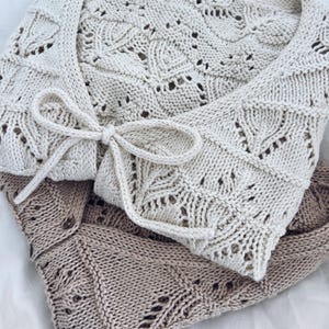 May include: Cream-colored knitted wrap with a delicate leaf pattern and a tie closure. The wrap is made of soft, textured yarn and is folded over a light brown knitted wrap. The wraps are displayed on a white surface.