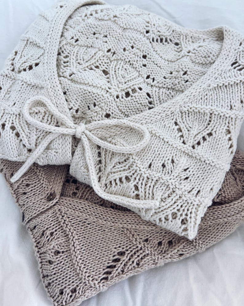 May include: Cream-colored knitted wrap with a delicate leaf pattern and a tie closure. The wrap is made of soft, textured yarn and is folded over a light brown knitted wrap. The wraps are displayed on a white surface.