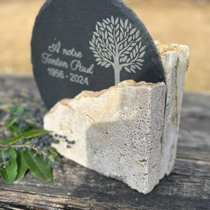 May include: A black slate memorial stone with the inscription "À notre Tonton Paul 1956-2024" and a tree design. The stone is held in a light-colored, textured stone holder, resting on a wooden surface.